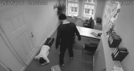 In einer nachgestellten Szene ist eine Büro-Situation zu sehen. Eine Mitarbeiterin liegt am Boden, über ihr steht ein Mann, der sie vermutlich niedergeschlagen hat. 