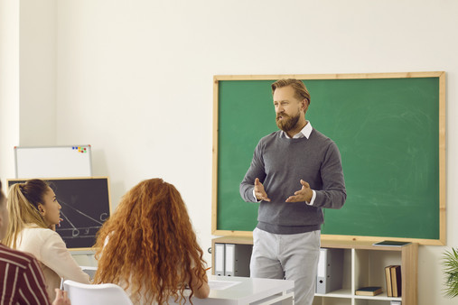 Ein Lehrer steht in einem Klassenzimmer vor der Tafel und spricht zu Schülerinnen und Schülern, die vor ihm an Tischen sitzen und zuhören. 