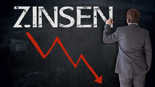 Zu sehen ist ein Mann, der mit Kreide "Zinsen" auf eine Tafel schreibt. Darunter ein roter Pfeil, der nach unten zeigt.