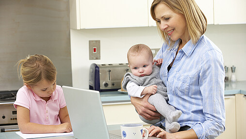 Foto zum Thema "Frauen im öffentlichen Dienst altersvorsorge": Zu sehen ist eine Mutter mit zwei Kindern in der Küche, die am Laptop arbeitet.