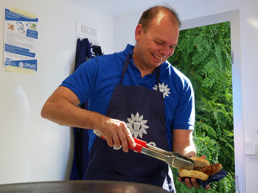 Reportage Einsatzversorgung - Daniel Heyer Foto zum Thema "Polizei Einsatzversorgung": Zu sehen ist Daniel Heyer am Grill. Er legt mit der Grillzange eine Wurst in ein Brötchen.