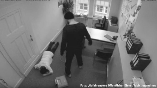 Symbolfoto der Kampagne "Gefahrenzone Öffentlicher Dienst": Das Bild einer Videokamera zeigt ein Büro, in dem ein Täter eine Mitarbeiterin scheinbar gerade zu Boden geworfen hat. 
