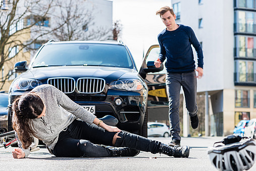 Eine Fahrradfahrerin wurde offensichtlich von einem Auto angefahren, sie liegt verletzt vor dem Auto auf der Straße. Der Fahrer des Wagens läuft auf sie zu. 