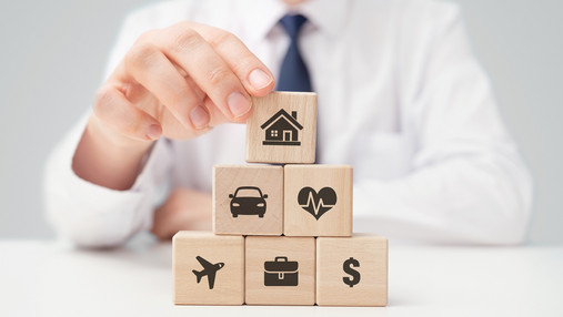 Was man nicht ausblenden sollte Das Foto zeigt die Hand eines Mannes, die eine Pyramide aus Bauklötzen aufstellt. Darauf sind Icons von verschiedenen Lebensbereichen abgedruckt, die man versichern kann.