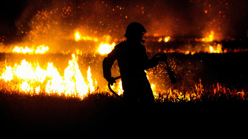 Foto zum Thema "Feuer um Wald": Zu sehen ist ein Feuerwehrmann, im Hintergrund lodern Flammen im Wald.