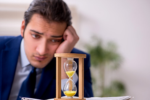 Junger Verwaltungsmitarbeiter sitzt am Schreibtisch und sieht verdrießlich auf eine ablaufende Sanduhr. 