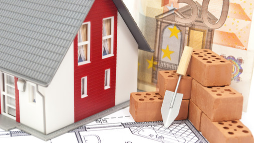 Zu sehen ist ein Modellhaus, daneben ein paar Bausteine, dahinter ein Geldschein. Das Foto ist ein Symbolbild für Immobilienfinanzierung.