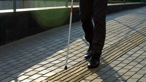 Blindheit - Leben mit einer Sehbehinderung Foto zum Thema Blindheit und Leben mit einer Sehbehinderung: Zu sehen ist eine blinde Person, die sich mit einem Blindenstock an einem Leitsystem orientiert.