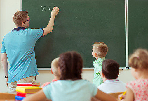 Junger Lehrer schreibt etwas an die Tafel, die Schüler*innen schauen zu. 