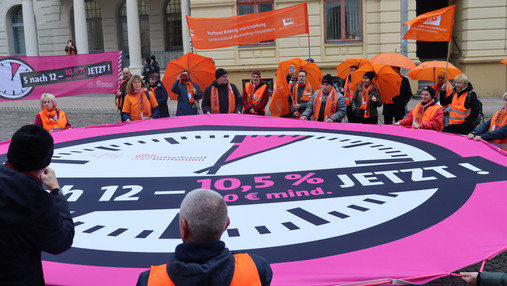 Demonstranten haben ein pinkes Banner in Form einer Uhr aufgespannt, auf dem "es ist 5 nach 12" geschrieben steht.