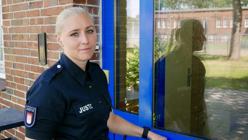 JVA Hahnöfersand - Dammann Foto zum Thema "JVA Hahnöfersand": Zu sehen ist Justizvollzugsbeamtin Dammann. Sie steht vor einer Tür und schaut in die Kamera.