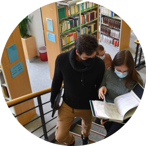 Studierende an der Hochschule des Bundes gehen in der Bibliothek eine Treppe herauf und sehen sich dabei ein Buch an. . 