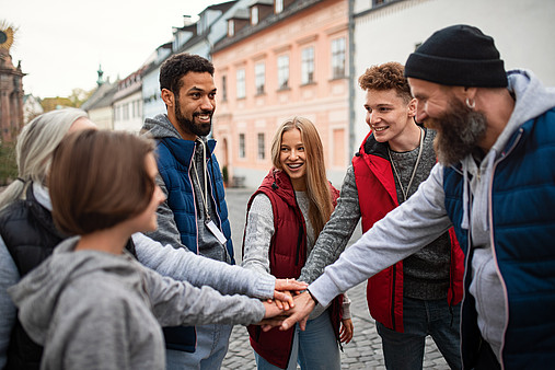 Eine Gruppe junger und älterer Menschen stehen im Kreis und haben die Hände als Zeichen des Zusammenhalts in der Mitte aufeinandergelegt. 