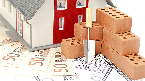 Foto zum Thema "Immobilienfinanzierung": Zu sehen ist ein Modellhaus, das 50-Euro-Banknoten steht. Davor Modell-Backsteine und eine kleine Schaufel.